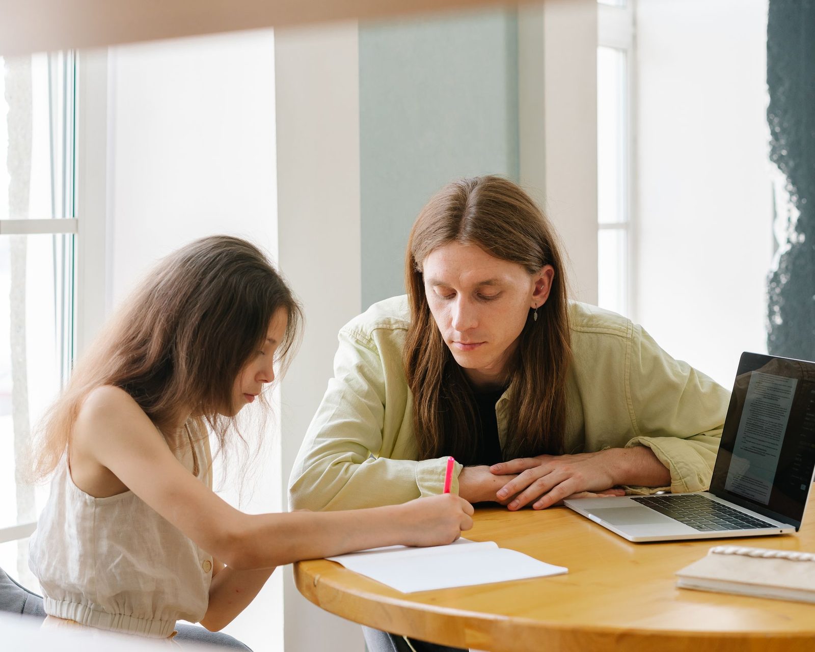 A parent and child working together at a sunlit table, writing in a notebook beside a laptop