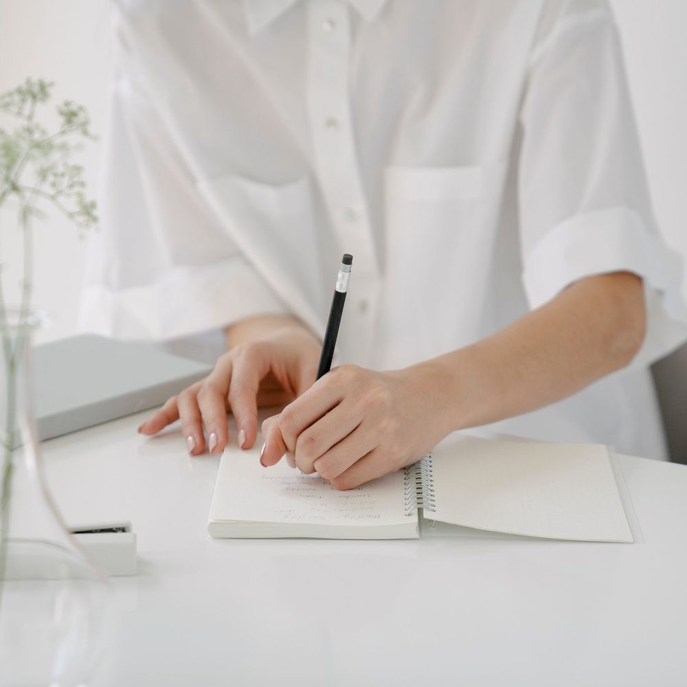 A person writing in a spiral notebook at a white table beside a vase of dried flowers